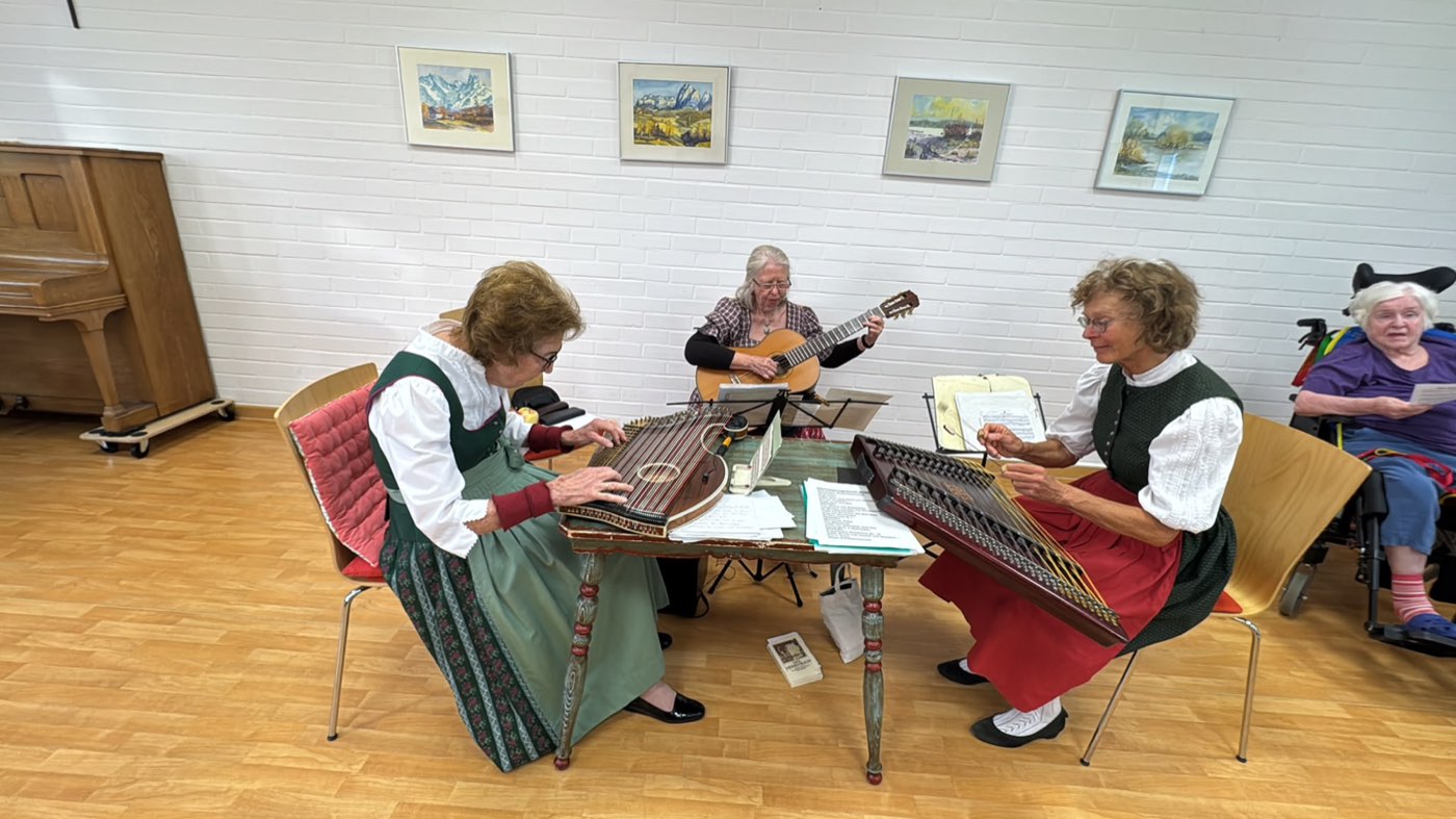 Drei Musikerinnen in Tracht spielen Zither und Gitarre, daneben hört ein Tagesgast zu